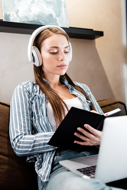 selective focus of attractive woman in wireless headphones writing in notebook near laptop in living room, online study concept