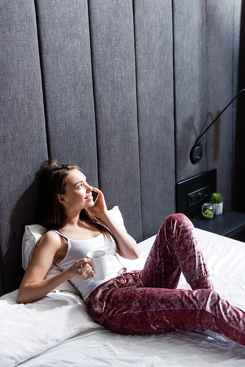 happy woman talking on smartphone and holding cup of coffee in bed