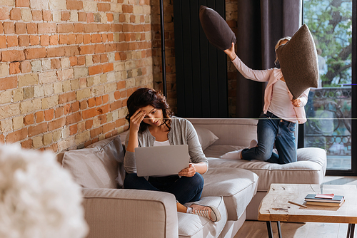 Selective focus of woman working on laptop near daughter playing pillow fight on couch