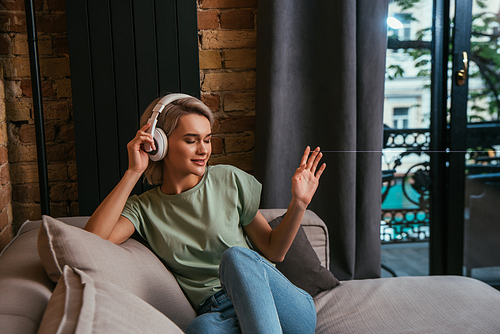 smiling woman gesturing while sitting on sofa and listening music in wireless headphones with closed eyes