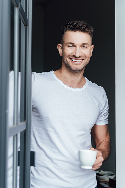 Selective focus of handsome man smiling at camera while holding cup of coffee near door at home