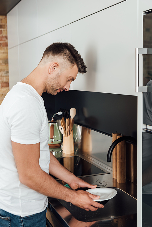 handsome man holding wet plate near faucet and sink in kitchen