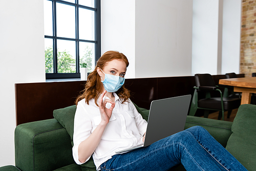 Woman in medical mask showing okay gesture while using laptop at home
