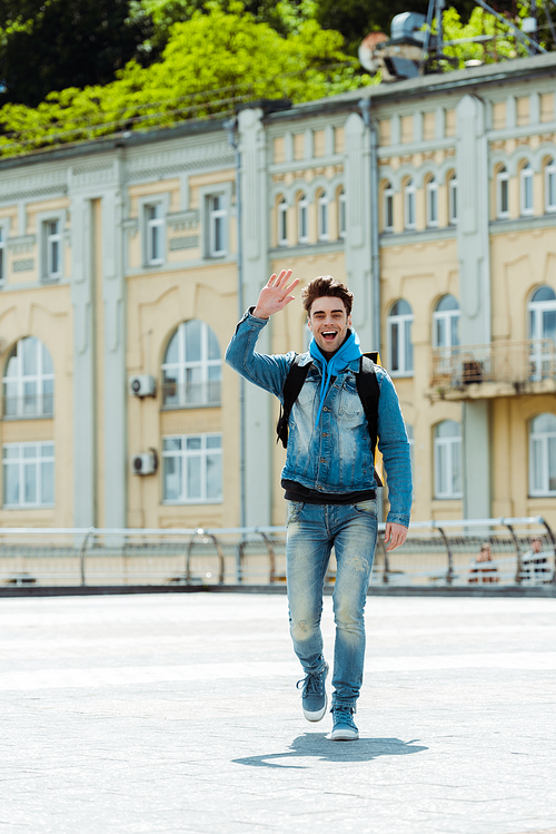 Cheerful delivery man with backpack waving hand on city street