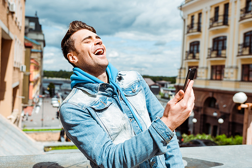 Handsome man laughing while holding smartphone on urban street