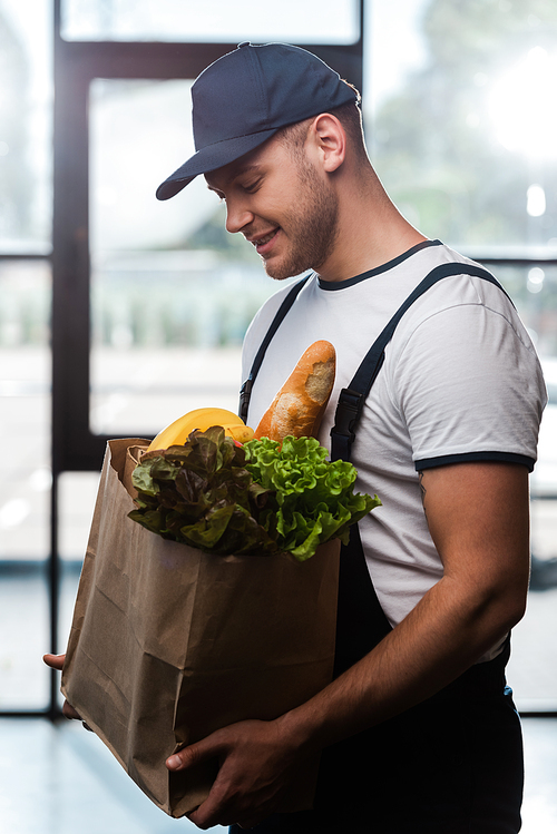 cheerful delivery man in cap looking at paper bag with groceries
