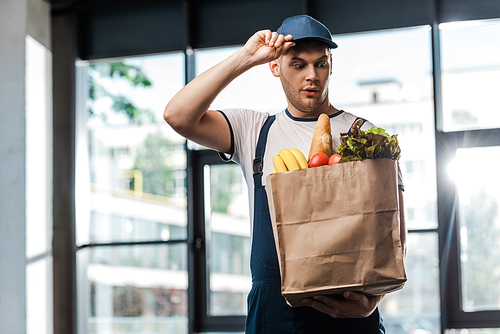 surprised delivery man touching cap and looking at paper bag with groceries
