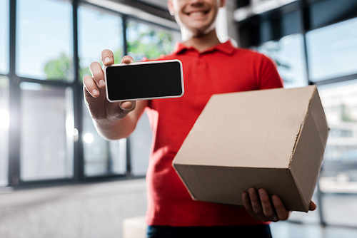 cropped view of happy delivery man holding smartphone with blank screen and carton box