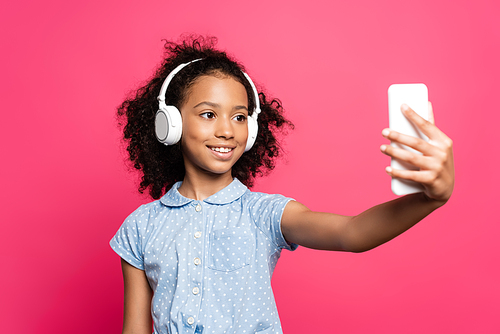 selective focus of smiling curly african american kid in headphones taking selfie on smartphone isolated on pink