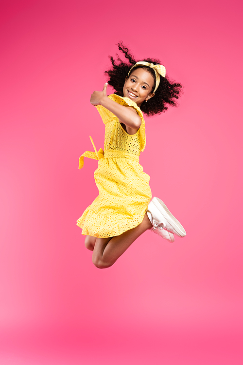 side view of happy curly african american child in yellow outfit jumping and showing thumb up on pink background