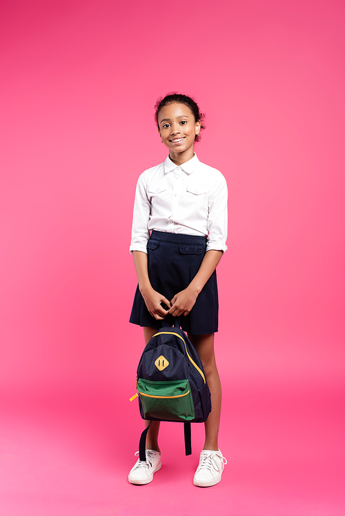 full length view of smiling african american schoolgirl with backpack on pink background