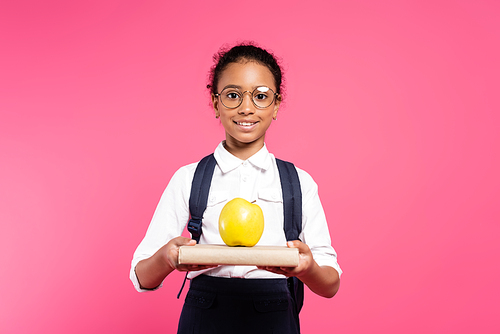 smiling african american schoolgirl in glasses with book and apple isolated on pink