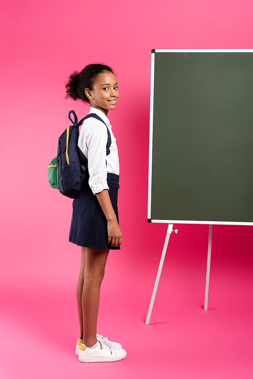 smiling african american schoolgirl with backpack near empty chalkboard on pink