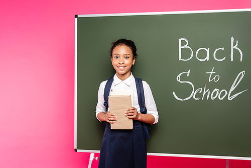 smiling african american schoolgirl with books near back to school inscription on green chalkboard on pink background
