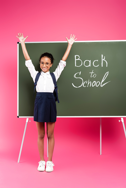happy african american schoolgirl in glasses with hands in air near back to school inscription on green chalkboard on pink background