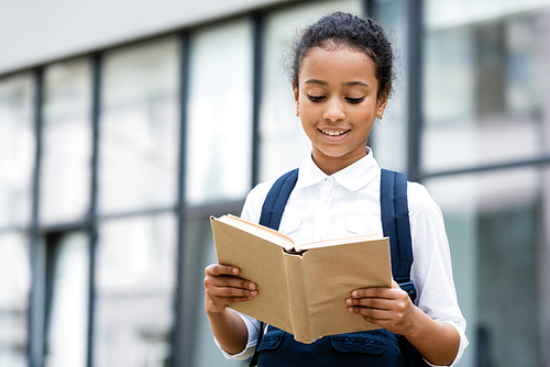 smiling african american schoolgirl reading book outdoors