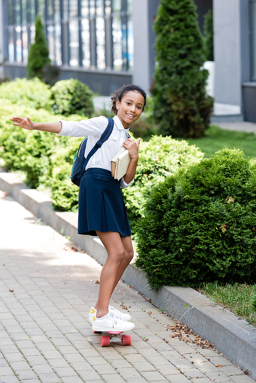 happy african american schoolgirl with backpack and books riding penny board outdoors