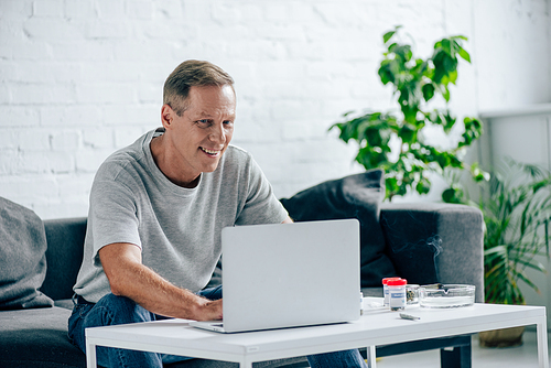handsome man in t-shirt smiling and using laptop in apartment