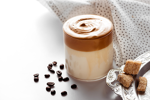 delicious Dalgona coffee in glass near coffee beans, brown sugar on silver platter and napkin on white background