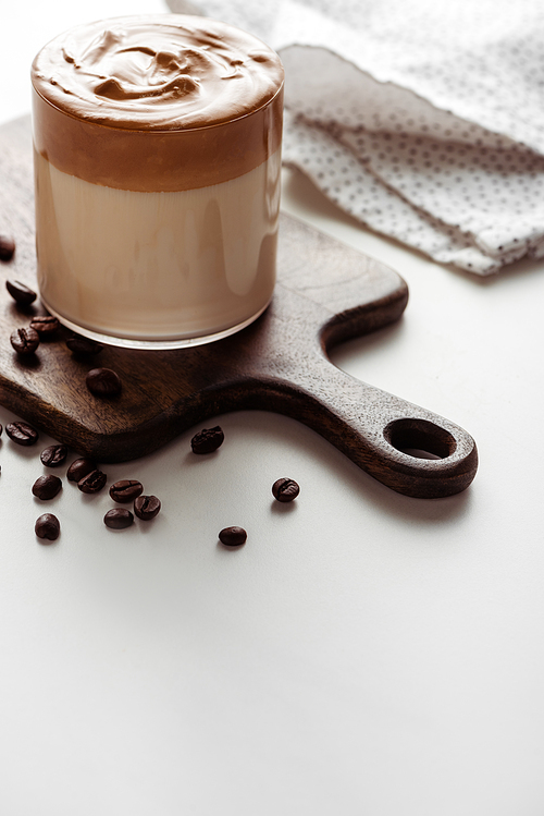 selective focus of delicious Dalgona coffee in glass on wooden cutting board near coffee beans on white background