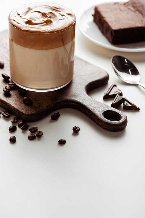 selective focus of delicious Dalgona coffee in glass on wooden cutting board near coffee beans, chocolate and spoon on white background
