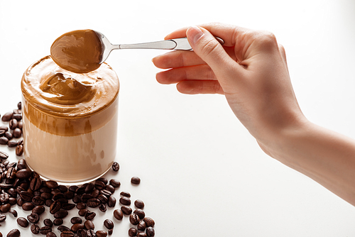 cropped view of woman holding spoon with foam near delicious Dalgona coffee in glass and coffee beans on white background