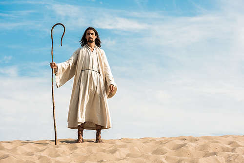 handsome man in jesus robe holding wooden cane against blue sky in desert