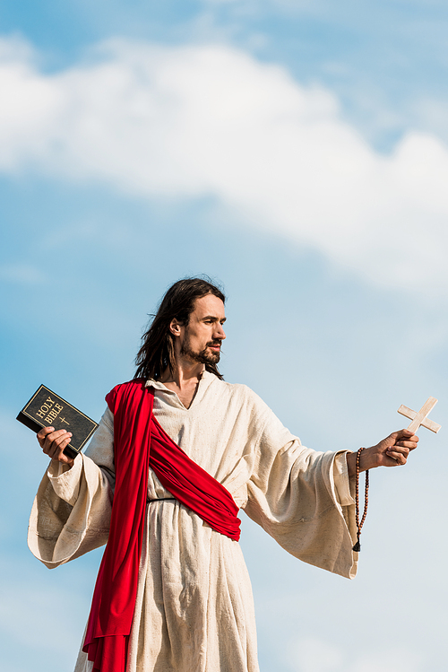 jesus holding holy bible and wooden cross against blue sky with clouds