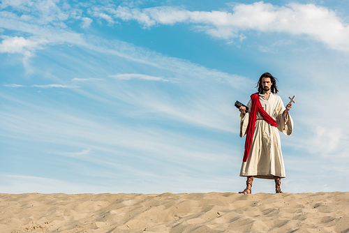 jesus holding holy bible and cross against blue sky with clouds in desert