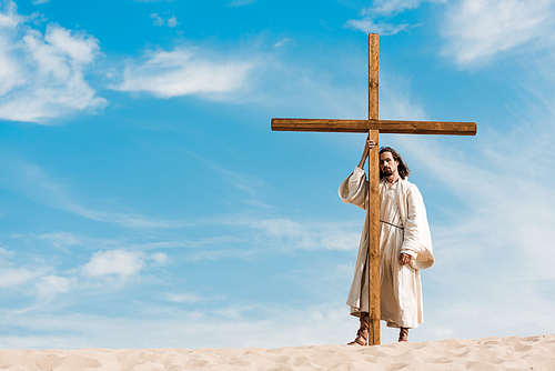 bearded man standing near wooden cross in desert on golden sand