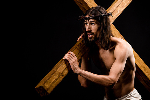 shirtless jesus in wreath holding heavy wooden cross isolated on black