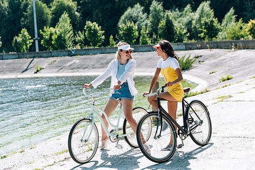 happy blonde and brunette girls with bikes looking at each other near river in summer