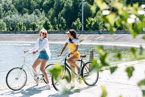 selective focus of green leaves and blonde and brunette girls riding bikes near river in summer