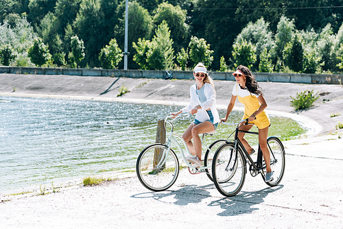 cheerful blonde and brunette girls riding bikes near river in summer