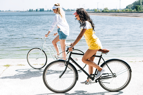 side view of smiling blonde and brunette girls riding bikes near river in summer