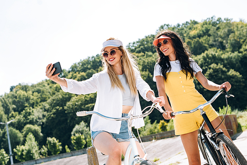 happy blonde and brunette girls riding bikes and taking selfie in summer