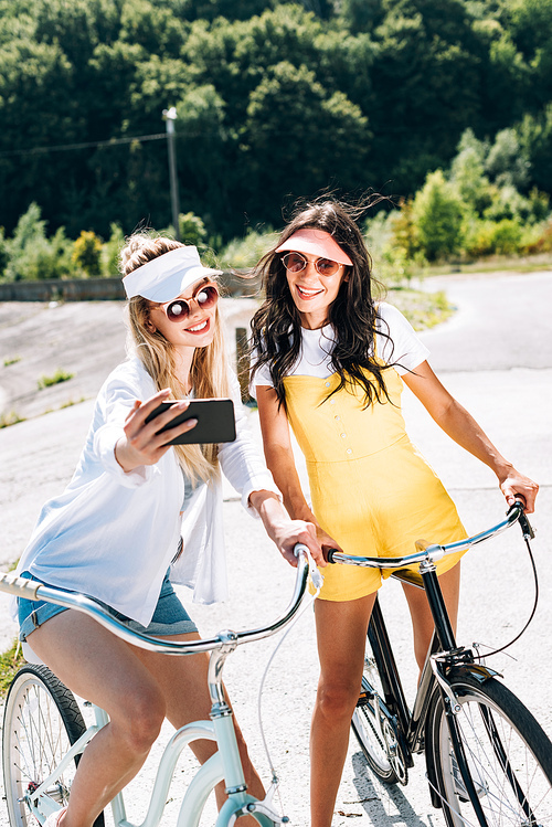 blonde and brunette girls riding bikes and taking selfie in summer