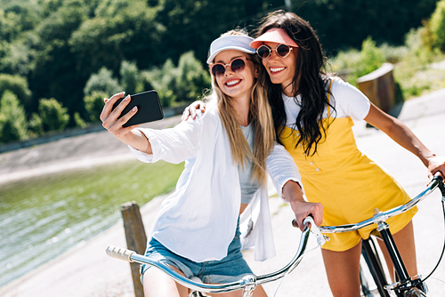 selective focus of blonde and brunette girls riding bikes and taking selfie in summer