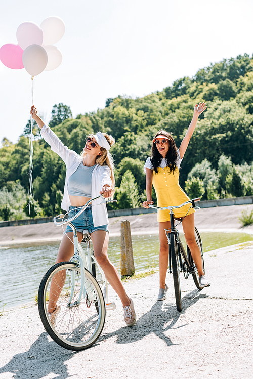 happy blonde and brunette friends riding bikes with balloons near river in summer