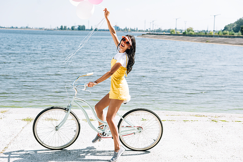 side view of happy brunette girl riding bike with balloons near river in summer