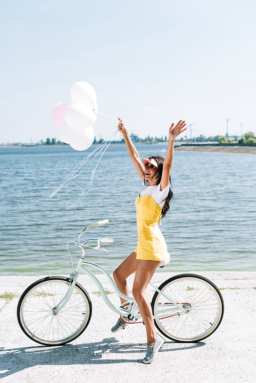 side view of happy brunette girl riding bike with balloons and hands in air near river in summer