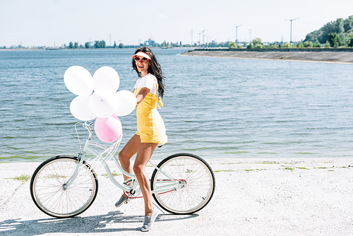 side view of beautiful brunette girl riding bike with balloons near river