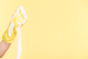 cropped view of cleaner in yellow rubber glove squeezing sponge with foam on yellow background