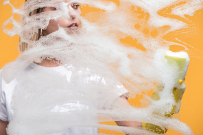 young woman in rubber glove with sponge cleaning glass covered with foam on orange background