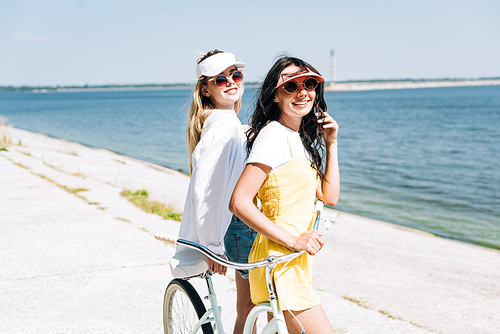 smiling blonde and brunette girls with bike near river in summer