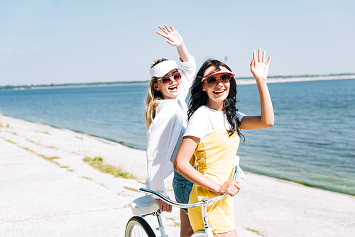 blonde and brunette girls with bike waving hands near river in summer