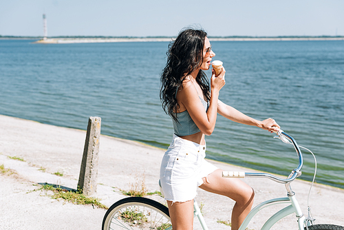 side view of brunette girl eating ice cream and riding bike near river in summer
