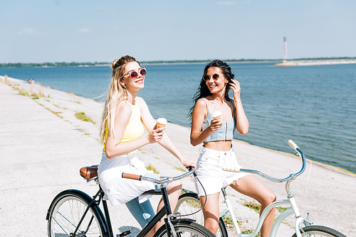 happy blonde and brunette girls riding bikes with ice cream near river in summer