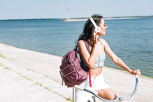 happy girl with backpack biking and listening music in headphones near river in summer