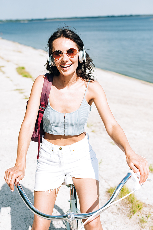 happy brunette girl biking and listening music in headphones near river in summer